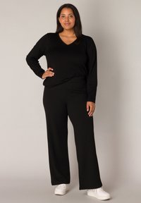 Zwarte langmouw V-hals top gecombineerd met brede zwarte broek. Materiaal lijkt zacht te zijn, met een gladde textuur. Gecombineerd met witte sneakers.