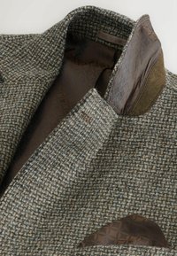 Jachetă din tweed în nuanțe mixte de maro și verde. Are o căptușeală maro cu model și un batic de buzunar din același material într-o formă triunghiulară.