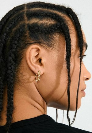 Goldener Ohrring in Form eines Hoops mit strukturierter Gestaltung und kleinen ornamentalen Akzenten. Haare in engen Zöpfen frisiert, mit feinen Details auf der Kopfhaut.