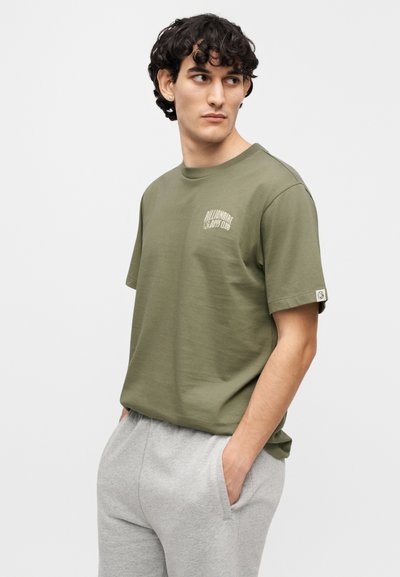 Μπλούζα από βαμβάκι σε ελιά πράσινο με κοντά μανίκια, στρογγυλή λαιμόκοψη και μικρό λογότυπο στο στήθος· συνδυασμένη με ανοιχτό γκρι παντελόνι φόρμας.