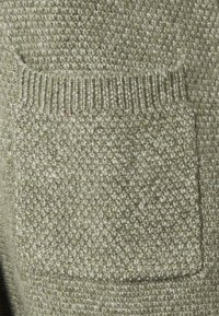 Tessuto a maglia verde oliva con una superficie testurizzata, dotato di una tasca a toppa e bordi a costine, indicativo di un design di abbigliamento informale.
