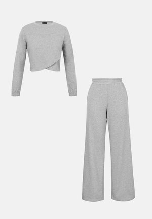 Ανοιχτό γκρι crop top με μακριά μανίκια και ασύμμετρο τελείωμα, συνοδευόμενο από παντελόνι με φαρδιά πόδια και ελαστική μέση, εμφανίζεται σε λευκό φόντο.