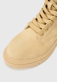 Gros plan sur une botte en suede beige avec de grosses lacets et une semelle robuste, mettant en valeur la texture et le savoir-faire.