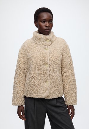 Femme portant une veste beige texturée boutonnée avec un col montant et un pantalon gris foncé, debout devant un fond blanc uni.