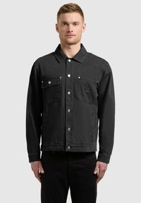 Man met kort haar, gekleed in een zwart jasje met knopen en twee borstzakken, en zwarte broek, met een effen achtergrond.