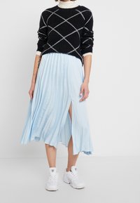 Zwarte trui met wit ruitpatroon, gecombineerd met een lichtblauwe geplooide midirok en witte sneakers. Zachte texturen en een gelaagd ontwerp.