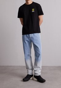 Černé bavlněné tričko s malou grafikou a textem v žluté, v kombinaci s světle modrými denimovými džínami, které blednou do bílé, a černými sportovními botami.