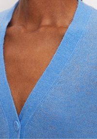 Hellblauer Cardigan mit V-Ausschnitt und einem einzelnen Knopf vorne, hergestellt aus weichem, strukturiertem Stoff mit feinem Strick.