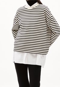 Proužkovaný pulovr v černé a krémové barvě s volným střihem a širokými rukávy, nošený přes bílou košili, v kombinaci s černými kalhotami.