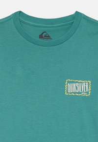Zila kreklu ar apaļu kaklu, kuram kreisajā augšējā krūtī ir neliels dzeltenu robežu "QUIKSILVER" logo.