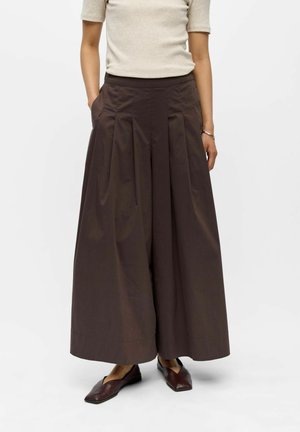 Femme portant un pantalon plissé marron foncé à jambes larges, un haut beige à manches courtes, des chaussures plates marron et un bracelet argenté, mains dans les poches.