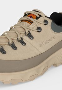 Μπεζ ορειβατικό παπούτσι Columbia με παχιά σόλα, μαύρες λεπτομέρειες, μεταλλικά γάντζους για κορδόνια και υφασμένη εξωτερική σόλα για πρόσφυση σε εξωτερικούς χώρους.
