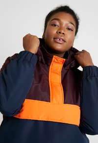 Chaqueta acolchada en azul marino y burdeos con una atrevida franja horizontal naranja. Presenta un alto cuello y puños elásticos para mayor comodidad.