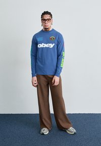 Camisa de manga larga azul con logo blanco de "obey", acentos verdes y rojos; pantalones anchos de cuadros marrones; zapatillas deportivas grises; gafas de estilo moderno.