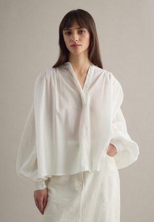 Mujer con cabello largo y castaño, vestida con una blusa blanca translúcida y holgada con mangas abullonadas y una falda blanca texturizada, de pie contra un fondo liso.