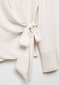 Svetle béžová wrap blůza s uvázaným předním dílem, vyrobená z hladké tkaniny. Má široké rukávy s ohrnutými manžetami a jemným leskem.
