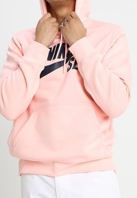 En person i en rosa Nike-hoodie som justerar huvan, bär silverringar och har en del av vita byxor synliga.