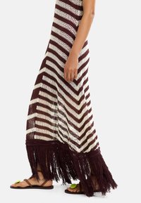 Szydełkowa maxi sukienka w bordowe i kremowe paski w zygzak, z długimi frędzlami na dole. Połączona z brązowymi sandałami i zielonymi akcentami.