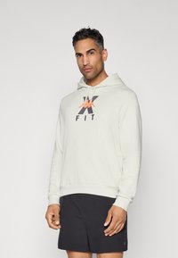 Mand med kort, mørkt hår iført en lysegrå hoodie med logo og sorte shorts, der står foran en ensfarvet lys baggrund.