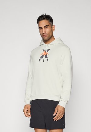 Man with short dark hair wearing a light hoodie with logo and black shorts, standing against a plain light background.