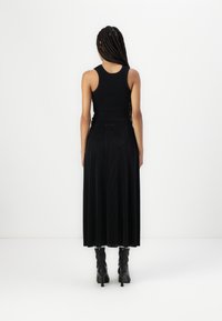 Haut sans manches noir avec un col rond, assorti à une longue jupe plissée noire. Le mannequin porte des bottes à talons noires montantes.
