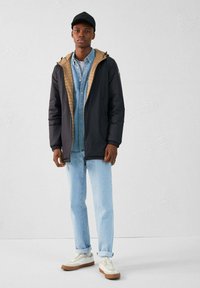 Chaqueta con capucha negra y marrón con interior acolchado, camisa de mezclilla azul claro, jeans azul claro remangados y zapatillas blancas con suelas marrones.