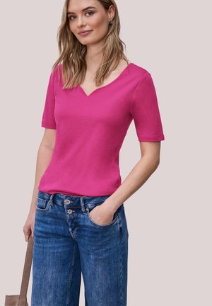 Frau mit glattem, blondem Haar, die ein hellrosa kurzärmliges V-Ausschnitt-Oberteil und blaue Jeans trägt, eine Hand in der Tasche, eine braune Tasche haltend.