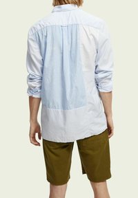 Camicia a maniche lunghe a righe verticali bianche e azzurre, con un pannello posteriore bianco e maniche con polsini, abbinata a pantaloni corti verde oliva.