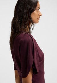 Donna con lunghe capigliature scure che indossa una blusa a maniche corte di un viola scuro con tessuto drappeggiato sulla spalla, rivolta verso destra su uno sfondo bianco.