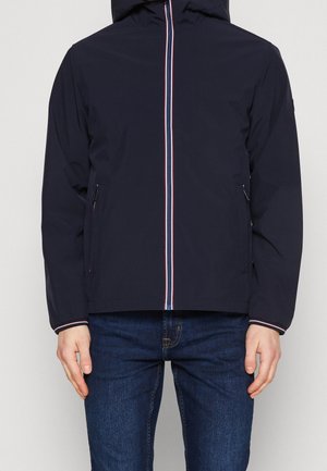 Mężczyzna ubrany w granatową kurtkę z kapturem, z czerwono-białym paskowanym zamkiem błyskawicznym i mankietami, w połączeniu z niebieskimi dżinsami, stoi na jednolitym tle.