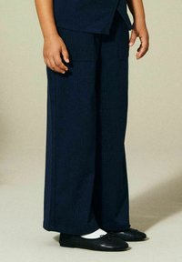 Barn står iført marineblå vide bukser, hvite sokker og svarte ballettsko foran en ensfarget beige bakgrunn.