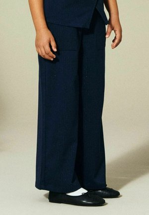 Bambino in piedi che indossa pantaloni navy a gamba larga, calzini bianchi e ballerine nere, davanti a uno sfondo beige uniforme.