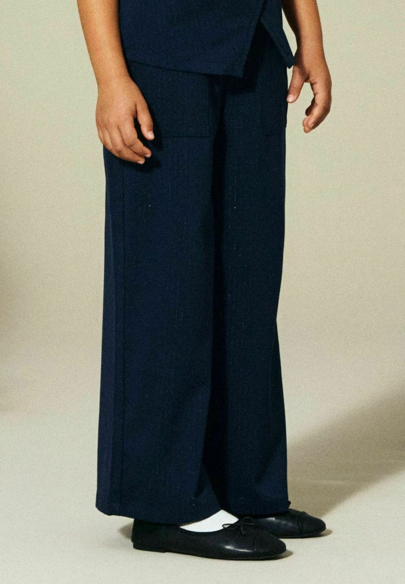 Barn står iført marineblå vide bukser, hvite sokker og svarte ballettsko foran en ensfarget beige bakgrunn.