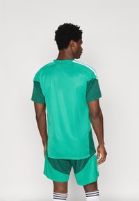 Camisola atlética verde com um painel lateral em verde escuro contrastante. Apresenta mangas curtas e riscas brancas de destaque. Usada com calções combinando.