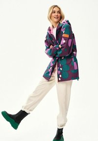 Kobieta uśmiechająca się, ubrana w białe spodnie, czarne buty z zielonymi podeszwami oraz fioletową kurtkę z dużymi zielonymi i różowymi okrągłymi wzorami.