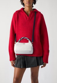 Sac à main blanc avec une finition texturée, présentant une anse torsadée en bleu et blanc. Porté sur un pull en tricot rouge et une jupe à carreaux.
