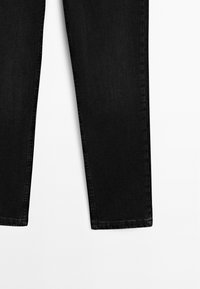 Partie inférieure d'un jean noir slim fit présenté sur un fond blanc montrant des ourlets droits et des coutures détaillées.