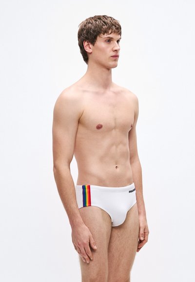 Weiße Badehose mit einem Regenbogenstreifen auf der linken Seite und einem schwarzen Logo am Bund. Glatte Textur, eng anliegend und minimalistisches Design.