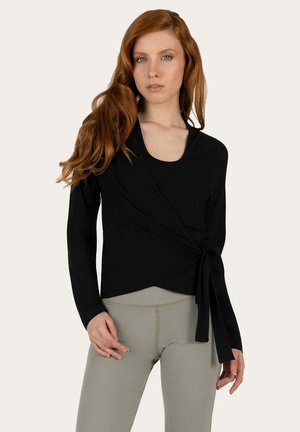 Femme aux longs cheveux roux portant un haut cache-cœur noir avec un lien sur le côté et des leggings taille haute gris clair, debout devant un fond neutre.