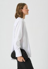 Valge pika varrukaga särk nööpide detailidega, koos tekstuurse musta käekotiga, millel on tikitud muster ja metallilised aktsendid.