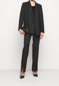 Schwarzer doppelt geknöpfter Blazer mit breiten Revers, kombiniert mit schmal geschnittenen Hosen mit seitlichen Schlitzen und einer kleinen schwarzen Clutch. High Heels runden das Outfit ab.