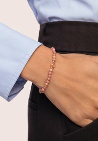 Vaaleanpunainen helminauha, jossa on pyöreitä kiviä vuorotellen kultakoristeiden kanssa, on käytössä ranteessa. Tausta on vaalea, ja näkyvissä on sininen paita.