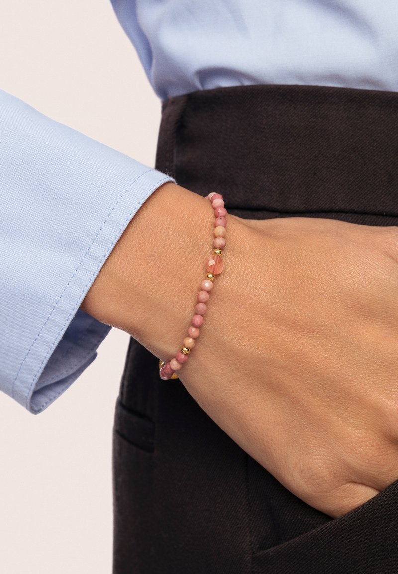 Vaaleanpunainen helminauha, jossa on pyöreitä kiviä vuorotellen kultakoristeiden kanssa, on käytössä ranteessa. Tausta on vaalea, ja näkyvissä on sininen paita.