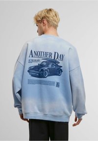 Vaaleansininen huppari, jossa on graafinen printti autosta ja tekstiä takapuolella. Kangas näyttää pehmeältä ja siinä on hieman haalistunut kuvio.