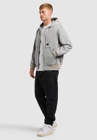 Bărbat tânăr în picioare, purtând o jachetă cu glugă de culoare gri deschis, tricou alb, blugi negri și adidași albi, pe un fundal simplu.