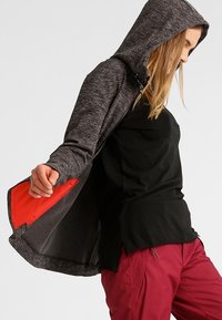 Szary hoodie o teksturowanym wykończeniu, z czerwonym wykończeniem i kapturem na sznurku, noszony na czarnej bluzce i bordowych spodniach.