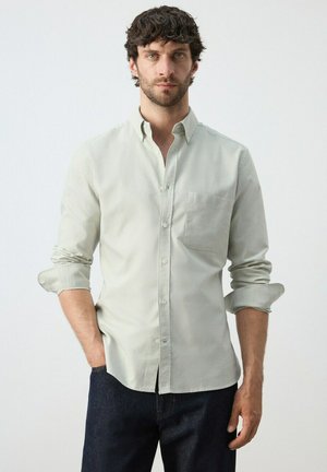 Hombre de cabello oscuro vestido con camisa gris claro de botones y mangas remangadas, y jeans oscuros, de pie contra un fondo blanco liso.