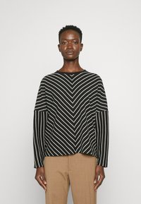 Black and white knitted sweater with chevron pattern, round neckline, and wide sleeves, paired with tan trousers. Textured fabric, casual fit.