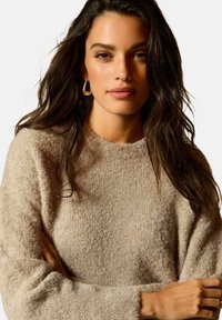 Jasno beżowy sweter wykonany z miękkiego, teksturowanego materiału, z okrągłym dekoltem i długimi rękawami. Modelka ma ciemne włosy i nosi kolczyki w kształcie kół.