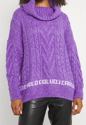 Kvinde iført en lilla kabelstrikket rullekravesweater med hvid tekst nær kanten, kombineret med sorte læderbukser.